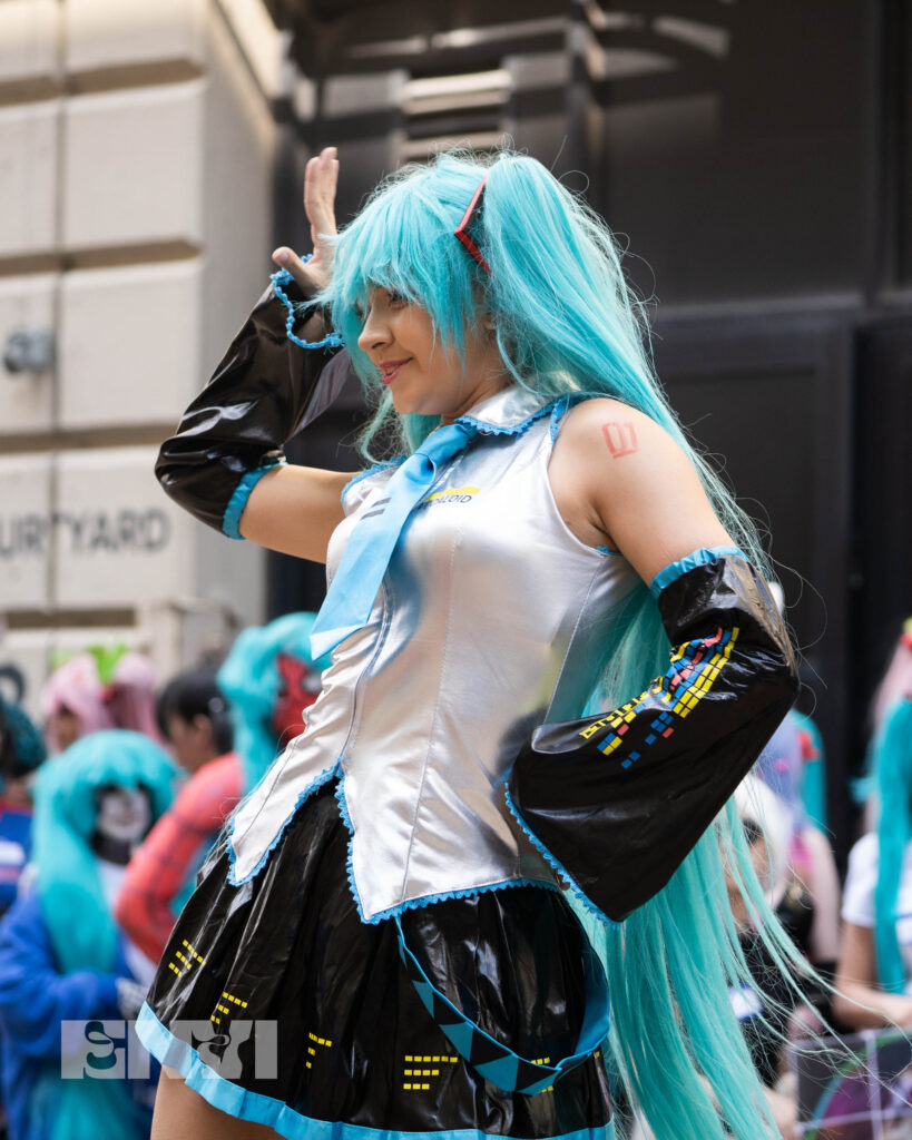 PearlHime is dressed as Hatsune Miku while performing to cosplayers at Japan Village. She holds one hand on her hip and the other by her eye.