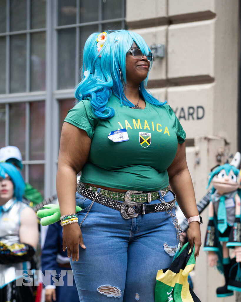 A cosplayer is dressed as a Jamaican Version of Hatsune Miku sporting a blue ombre wig and a shirt that prints "Jamaica" in all caps