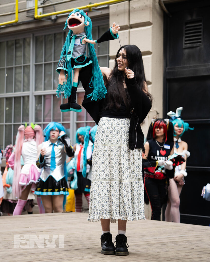 A competitor holds a Muppet version of Hatsune Miku in her hands. The Miku Character has Yarn Hair, and wears Black, Gray and Blue