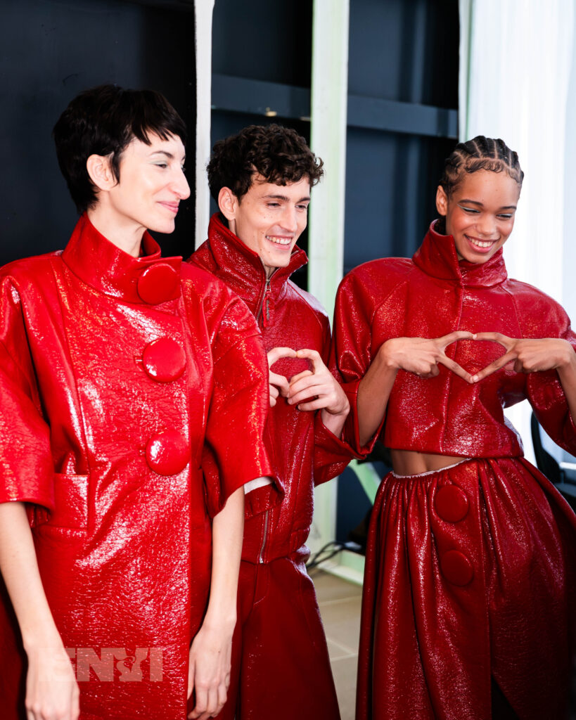 Three models in shiny red leather-looking jackets smile in a dressing room. Two of them make diamond shapes with their fingers.