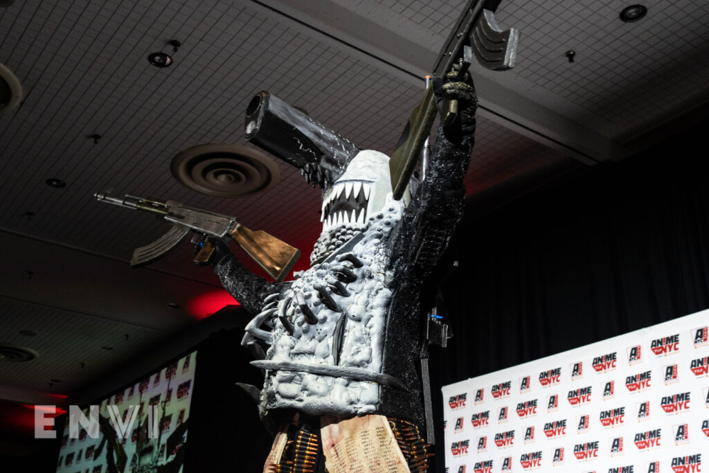 A cosplayer wears a white and black foam outfit with guns placed atop their head and arms.