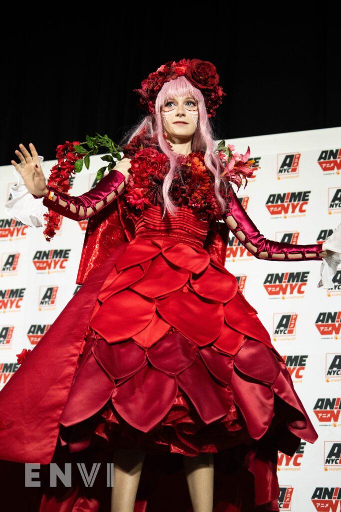 Mad Dog VA wears an ornate red satin gown as their arms are slightly extended. Wearing a pink wig, they look out to the crowd.