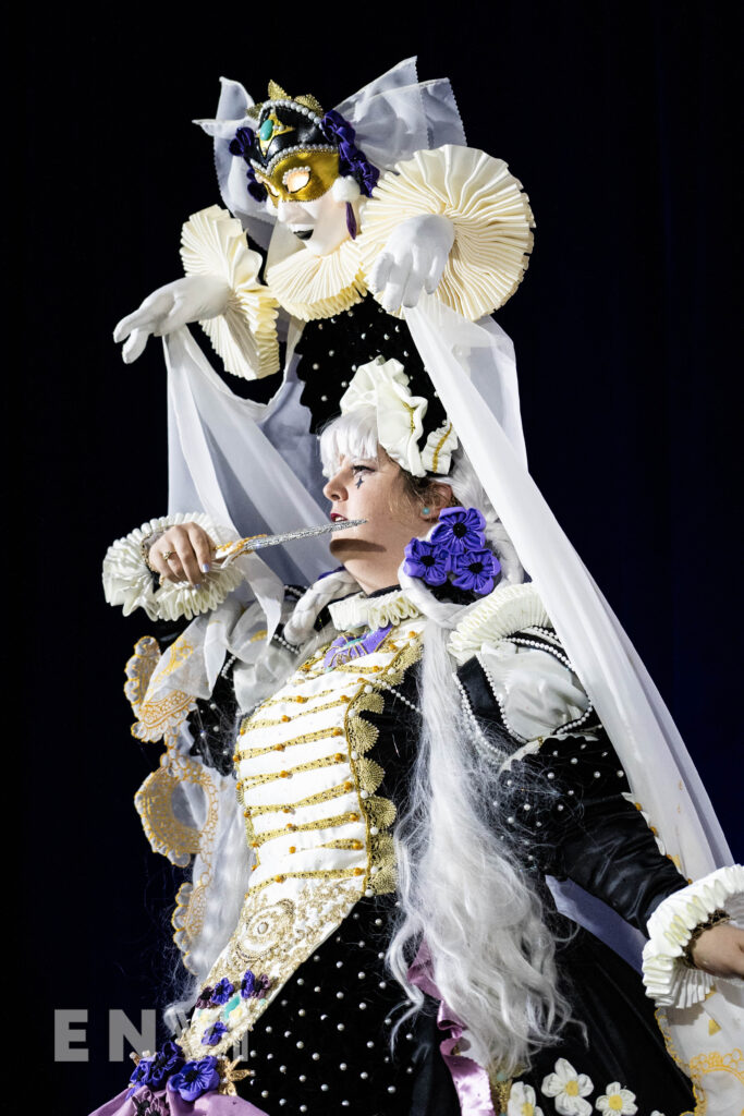 MissMina2 looks out to the crowd at the Anime NYC Cosplay Masquerade. The wear an ornate gown embellished with gold and purple trim.