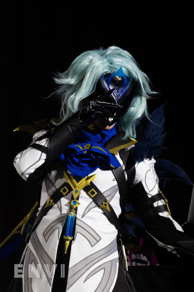 A cosplayer makes their way across the stage while holding their mask. They sport a blue wig with ornate gold and black trims.