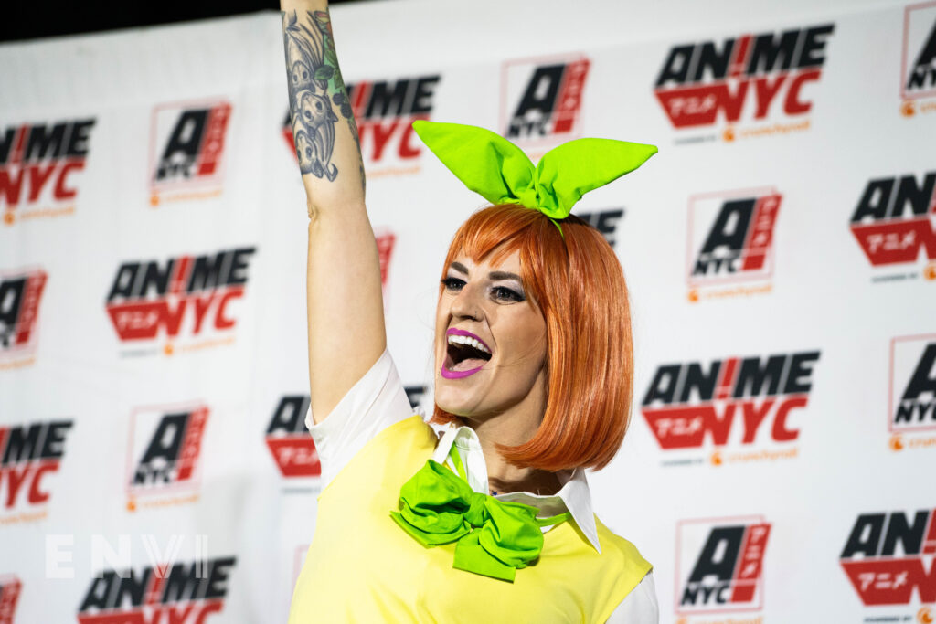 The Shaky Cosplayer wears an orange wig and yellow school uniform while smiling at the crowd and raising her right fist in the air