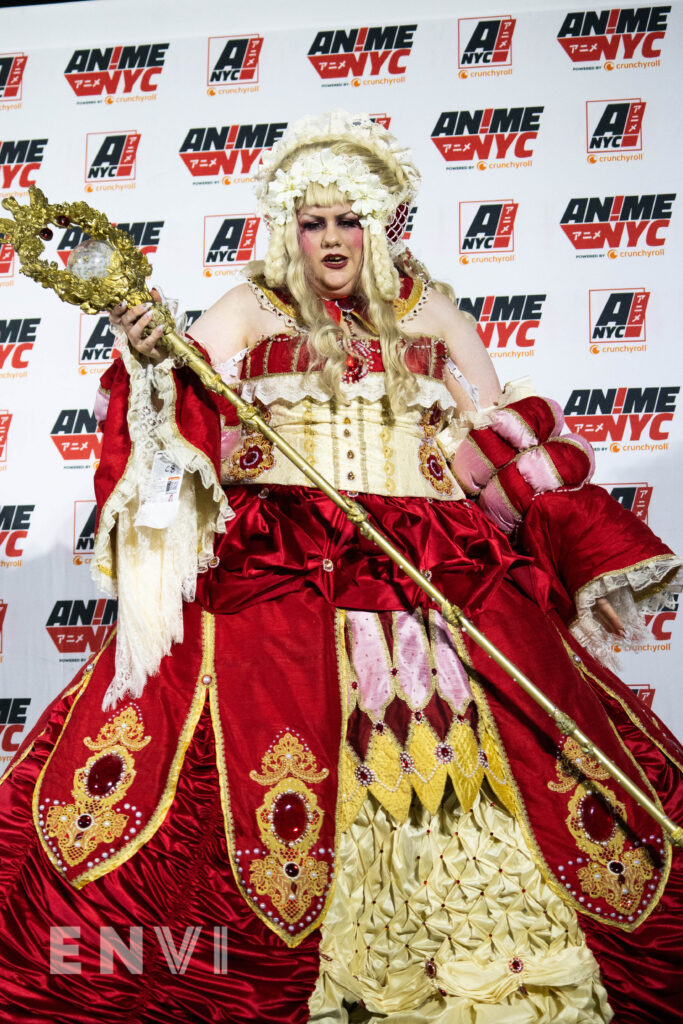 White Rabbit Cosplays looks off toward the camera holding an ornate gold staff and red gown.