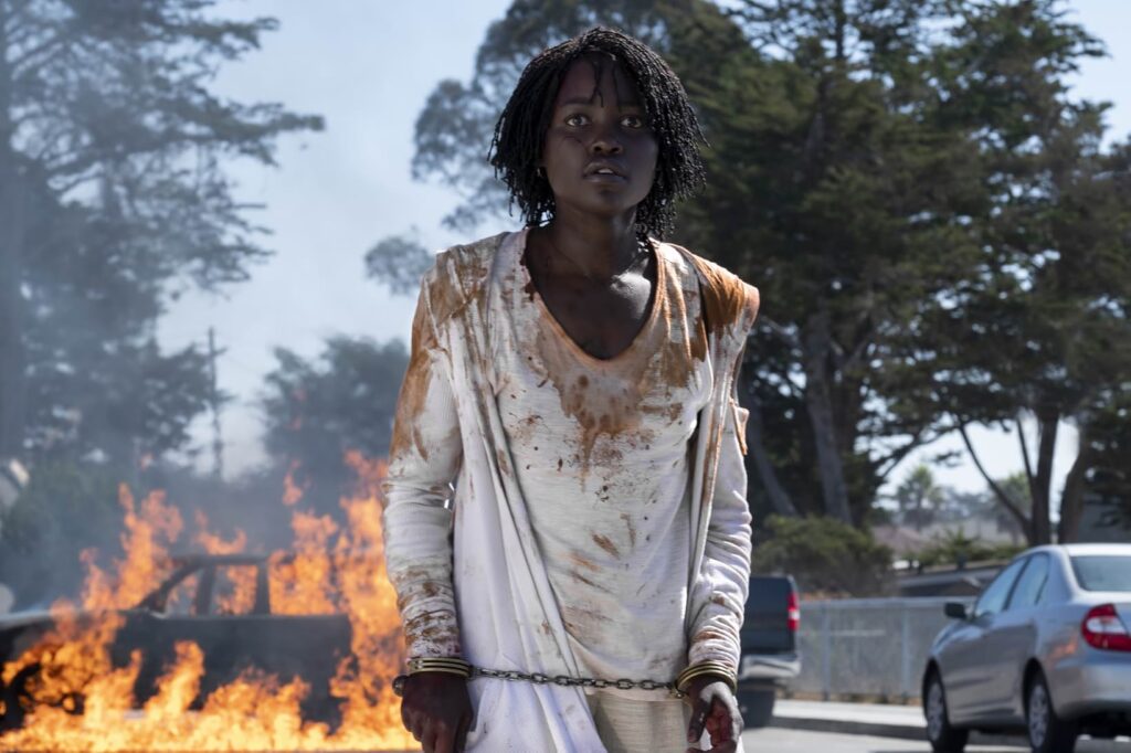 A young woman in chains and a stained white dress looks up while walking away from a car on fire