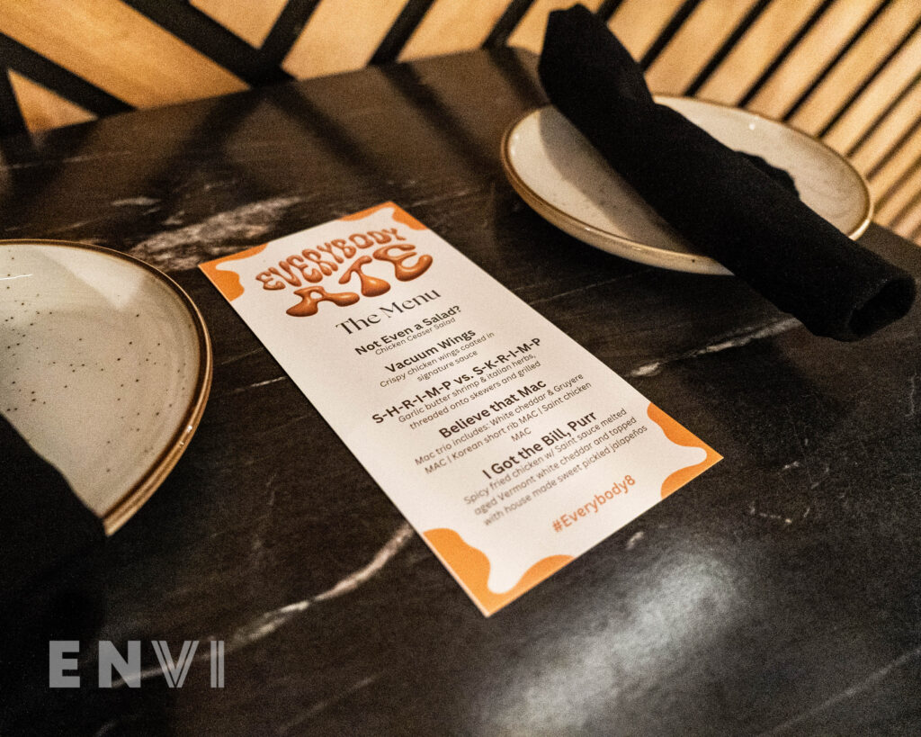 Two plates sit atop a table with Black napkins holding cutlery. In the middle of the table is a white and pink menu with Cleotrapa's branding.