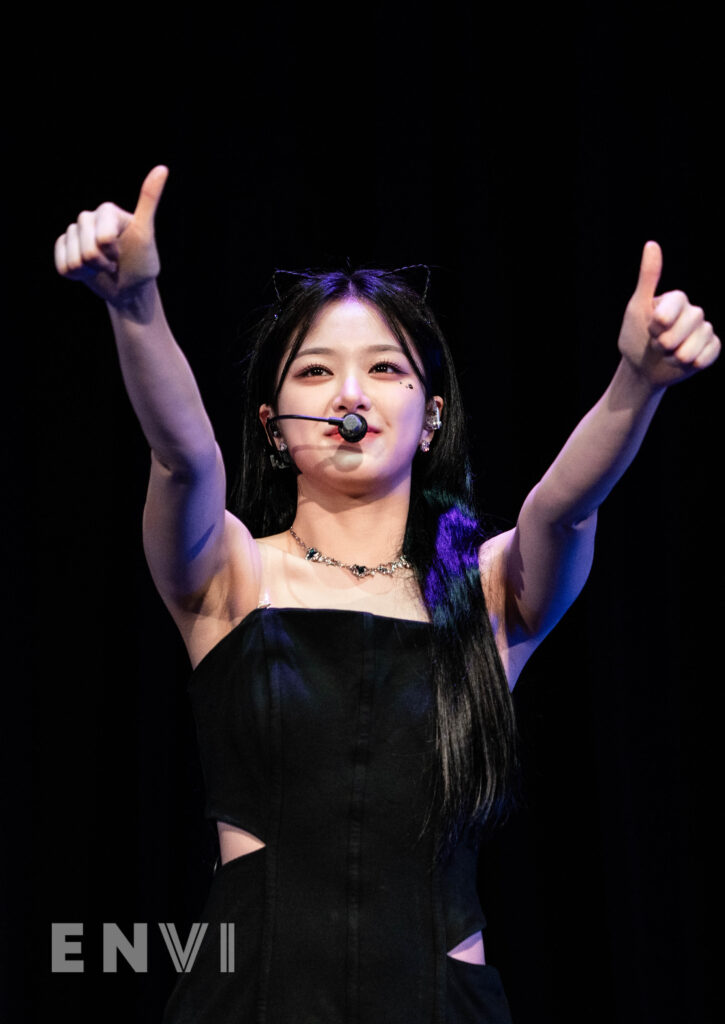 Hyunjin gives a thumbs up to fans with a smile on her face. She wears a black fitted dress and a necklace.