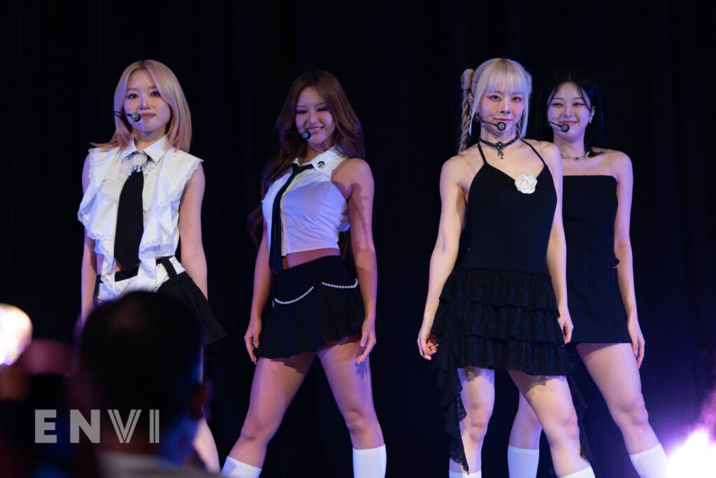 The members of Loossemble pose for fans at their concert at town hall. Pictured from left to right are members Gowon, Hyeju, Vivi, and Hyunjin. They wear a mix of black dressed and two piece skirt and button down outfits.