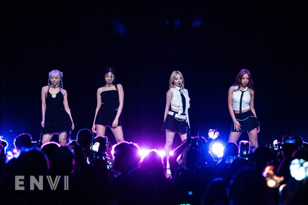 The members of Loossemble pose for fans at their concert at town hall. Pictured from left to right are members Vivi, Hyunjin, Gowon, and Hyeju. They wear a mix of black dressed and two piece skirt and button down outfits.