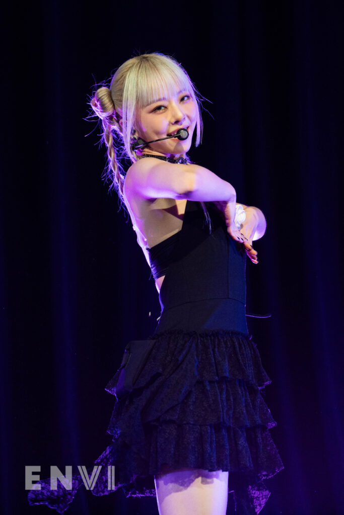 Vivi wears a black dress with blonde hair. She smiles as she holds her hands by her chest.