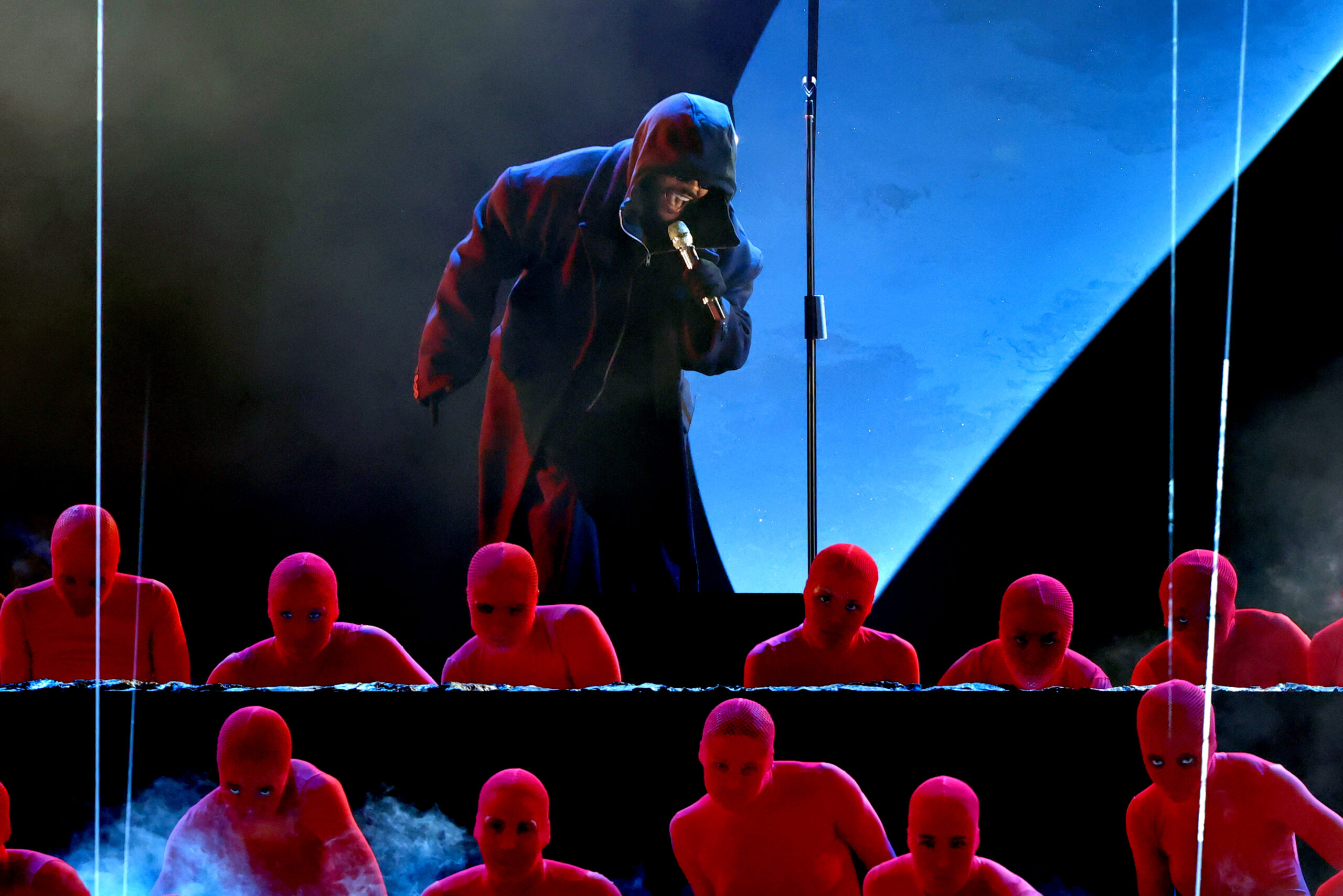 The Weeknd stands on stage performing at the 67th Annual Grammys holding a microphone in his hand. Wearing a hooded coat, he is surrounded by dancers dressed in red bodysuits.