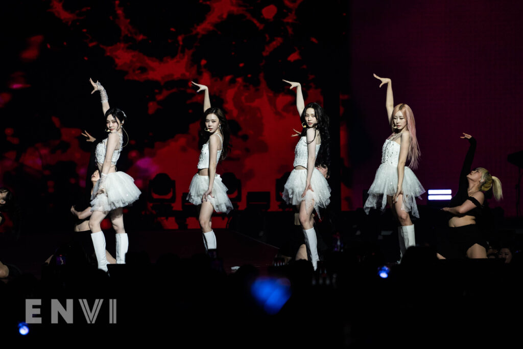 The members of K-pop girl group aespa end their performance of "Black Mamba" wearing white outfits embellished with rhinestones and jewels. They are posed with their bodies facing the sides and right arms lifted. Members are pictured from left to right: NINGNING, WINTER, KARINA, and GISELLE