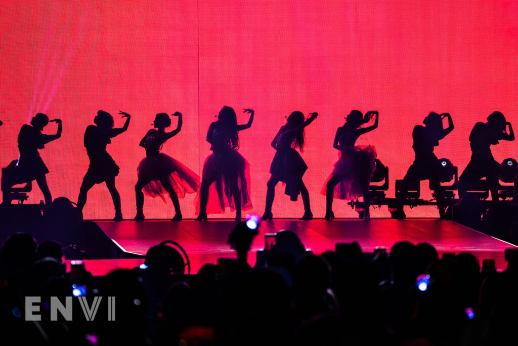 The members of aespa are joined by backup dancers who hold their left arms by their heads. They are in Silhouette as the red background of the screen flashes bright.