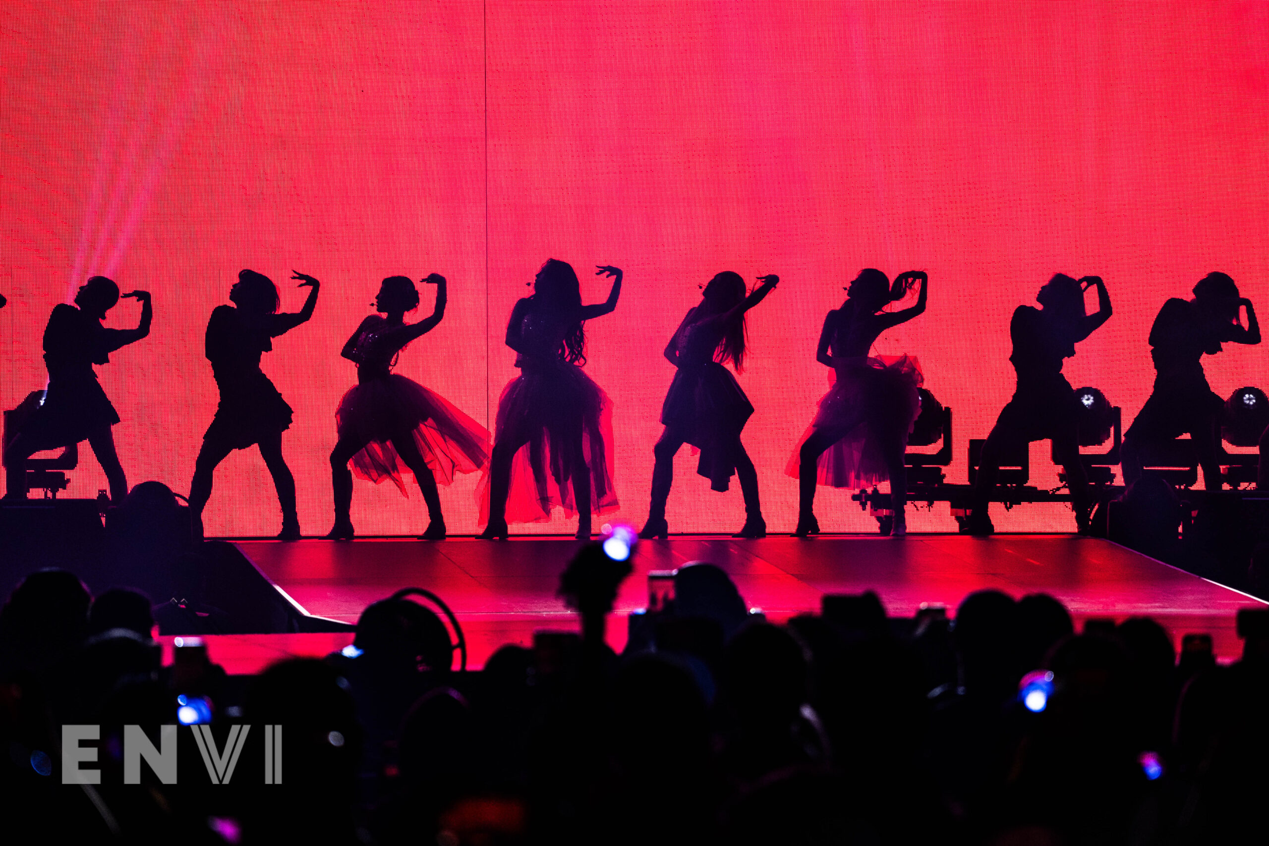 The members of aespa are joined by backup dancers who hold their left arms by their heads. They are in Silhouette as the red background of the screen flashes bright.