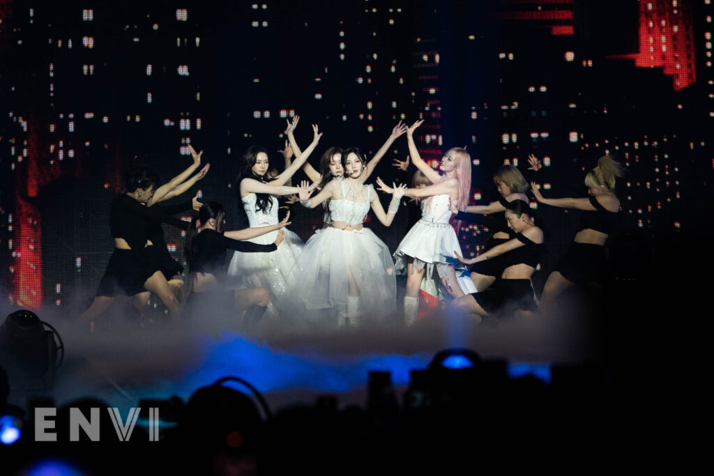 aespa dressed In all white perform their track "Drama." Surrounded by smoke and lights, dancers surround them with arms extended towards aespa.