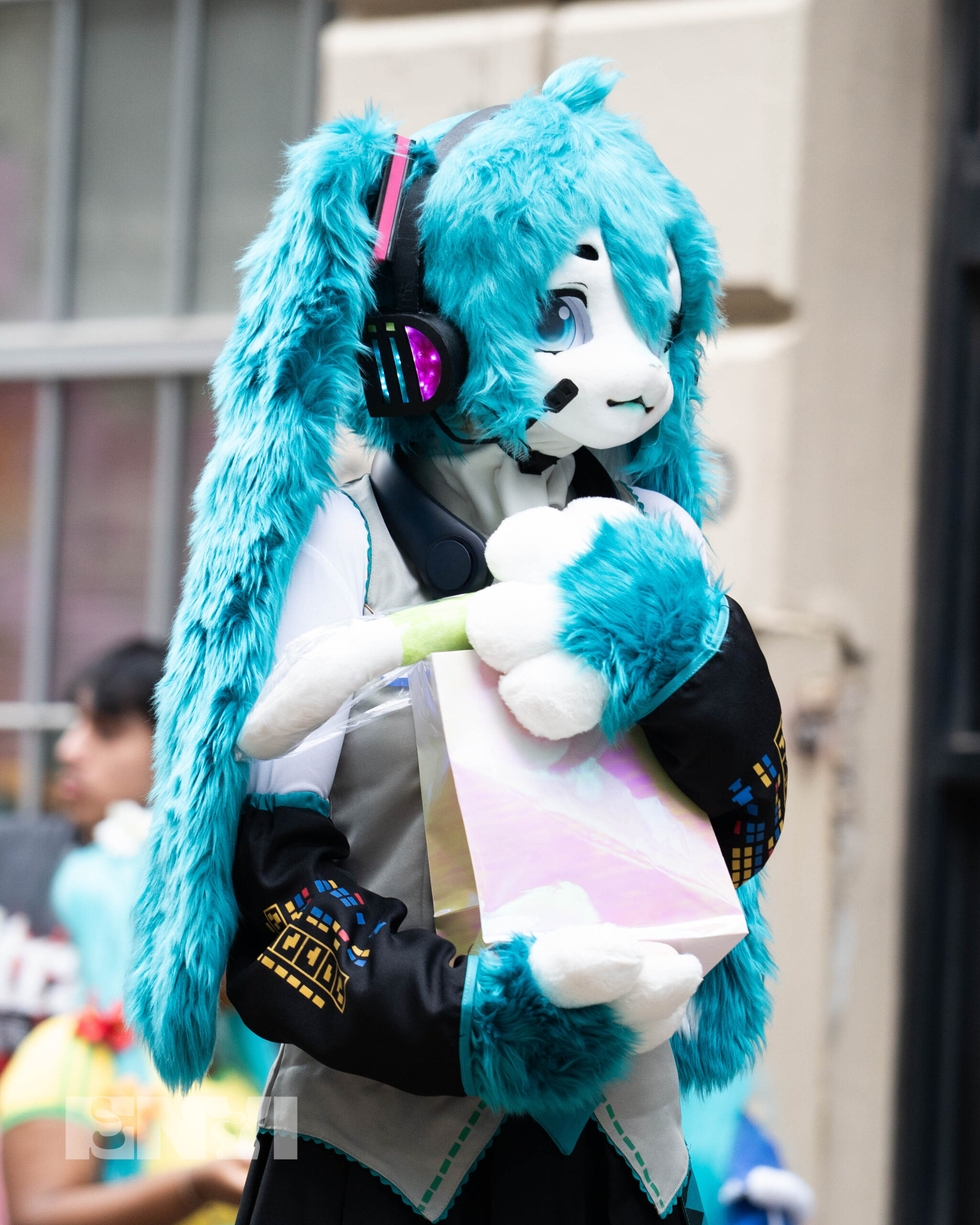 A competitor is dressed as a "Furry" version of Hatsune Miku wearing a full Fur suit with ears mimicking two long blue Pigtails. The cosplayer also holds a gift bag following their win at the contest