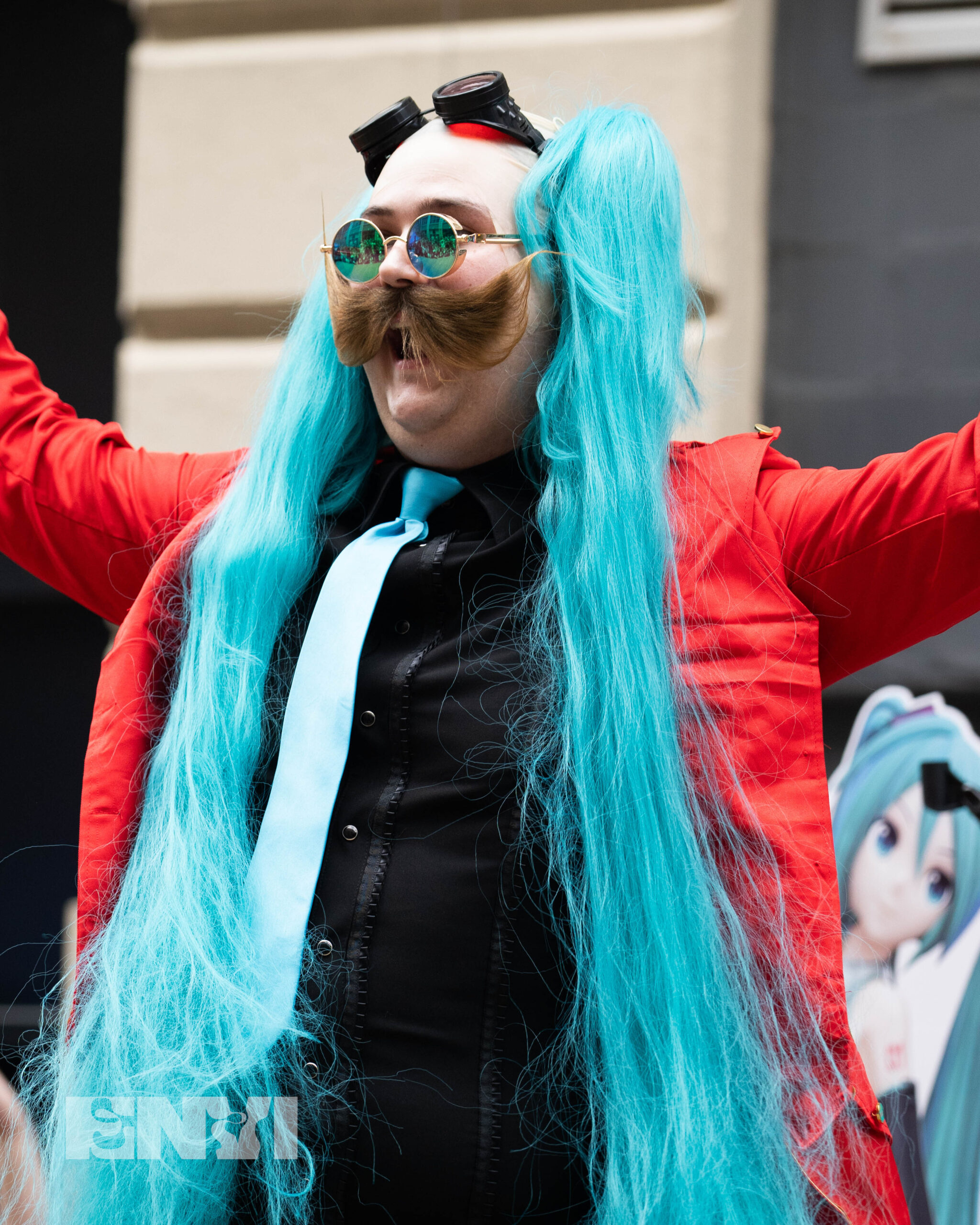 A cosplayer is dressed as a Eggman version of Hatsune Miku, mixing the Sonic Universe with the popular vocalic. They wear blue and gold detailed sunglasses with another set of goggles atop their head. Wearing a bald cap, two pigtails are fixed near their temples with a red suit and Black shirt.