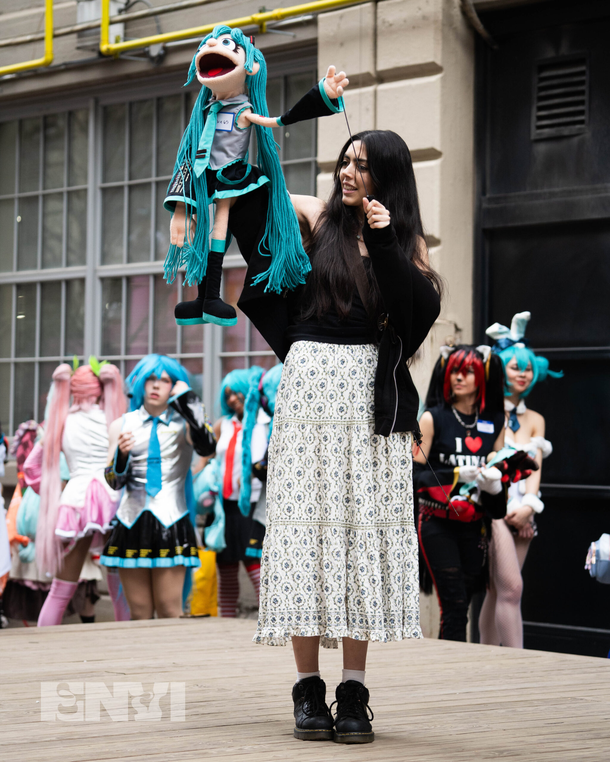 A competitor holds a Muppet version of Hatsune Miku in her hands. The Miku Character has Yarn Hair, and wears Black, Gray and Blue