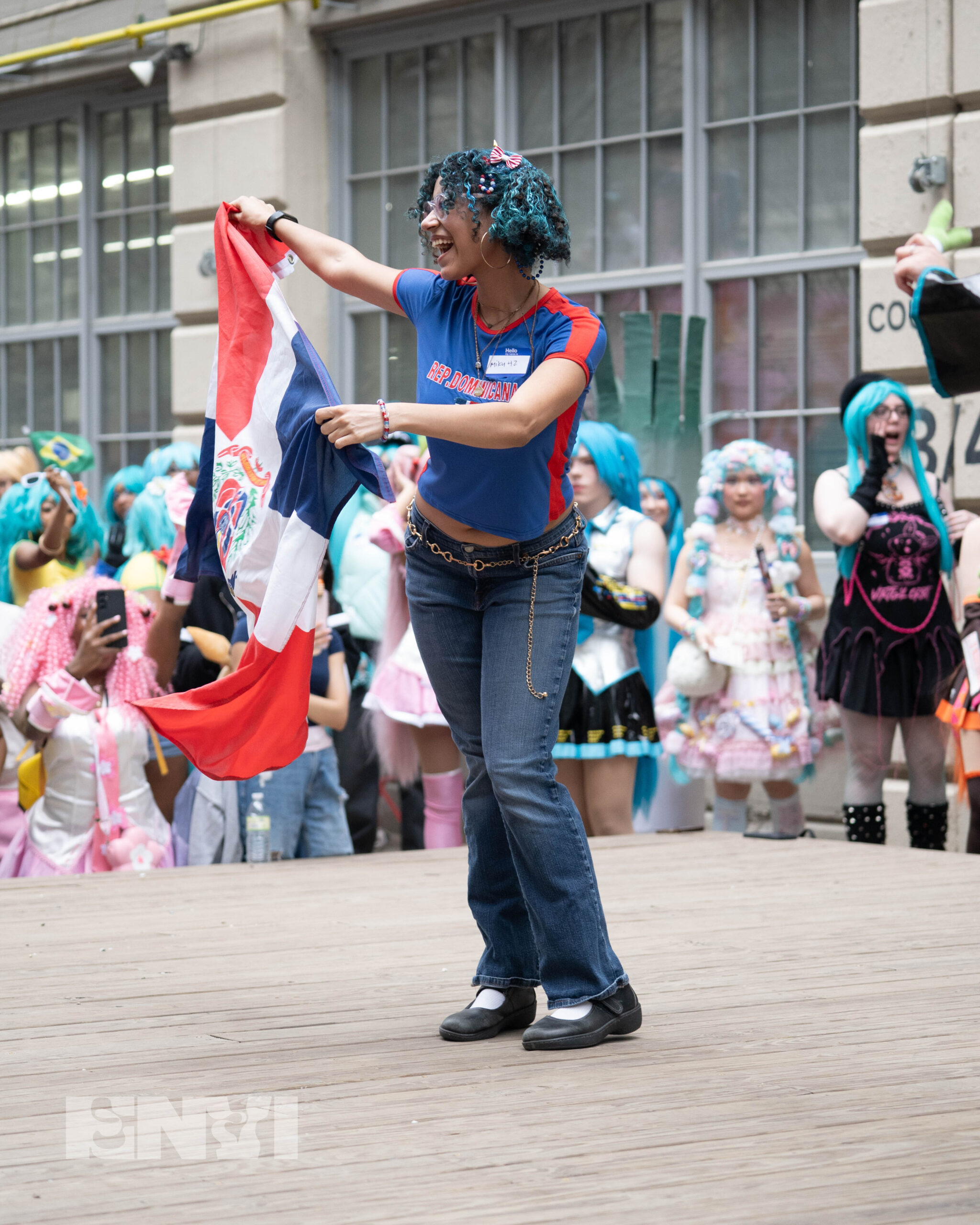 Araly is dressed as a Dominican version of Hatsune Miku wearing navy blue denim jeans, Mary Janes, and a red and blue fitted tee. Holding a Dominican Republic flag In their hand, they wave it with pride while wearing two blue pigtails.