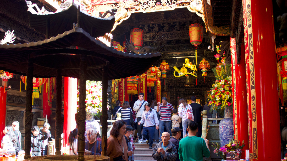 In the center of glossy red pillars and gold decorations is a standing censer with incense sticks. People bustle in and out of a temple doorway.