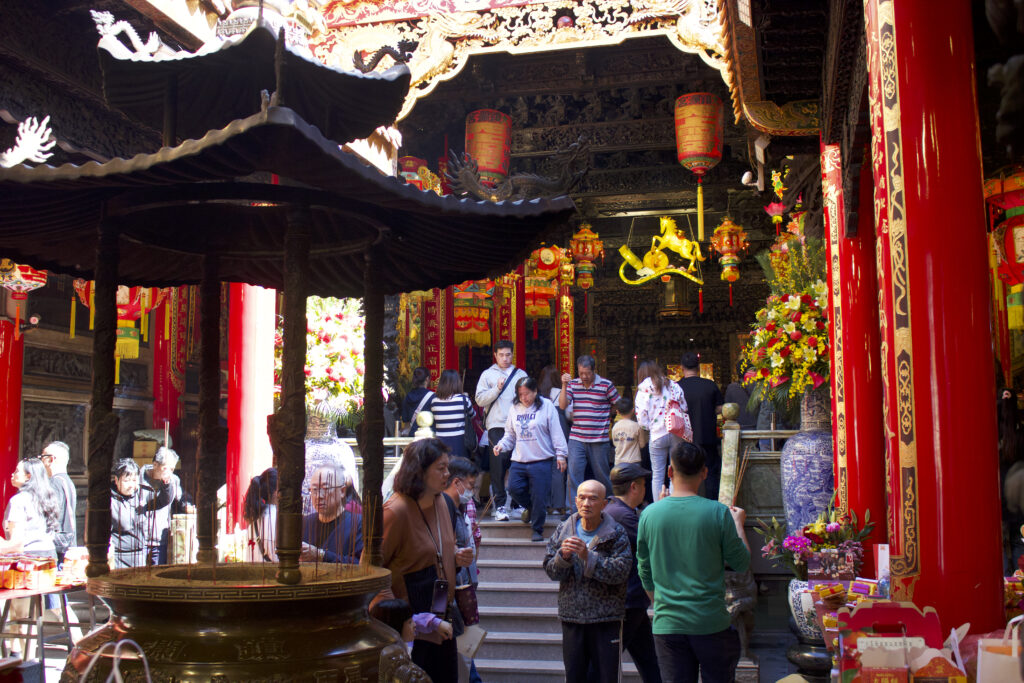 In the center of glossy red pillars and gold decorations is a standing censer with incense sticks. People bustle in and out of a temple doorway.