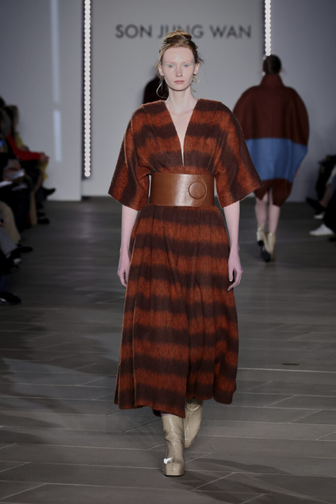A model on a runway wears a long loose dress striped with brown and burnt orange with a brown leather belt. She has bright green eyeshadow and pale cream ankle boots.