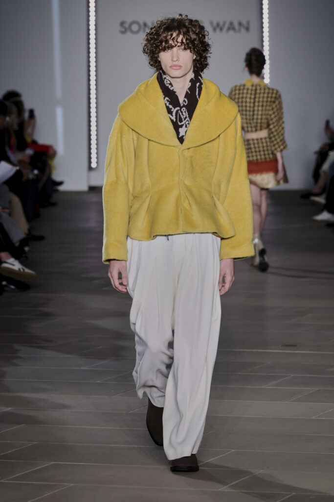 A model on the runway wears a fuzzy yellow coat with a brown scarf with white detailing underneath and loose white pants. 