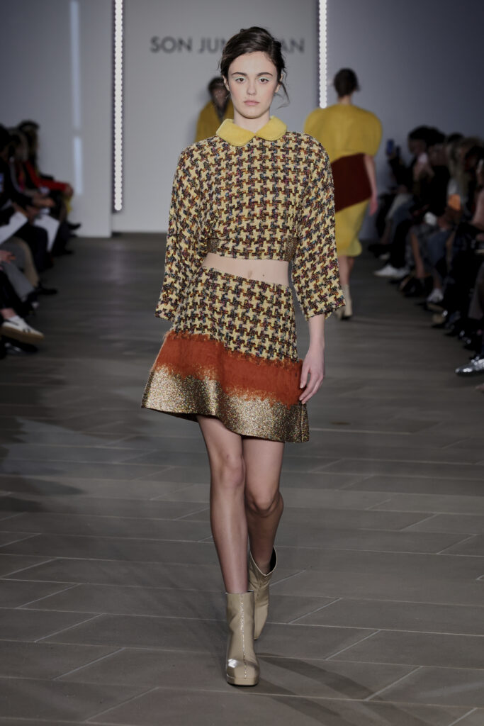 A model on a runway wears a yellow and brown tweed top and matching skirt. The skirt fades into burnt orange and metallic gold at the bottom.