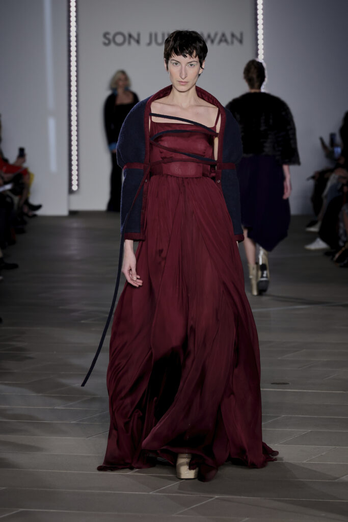 A model on the runway wears a burgundy flowing jumpsuit with a navy shawl with burgundy lining and edges. Her chest is crossed with straps over the tighter bodice of the jumpsuit. 