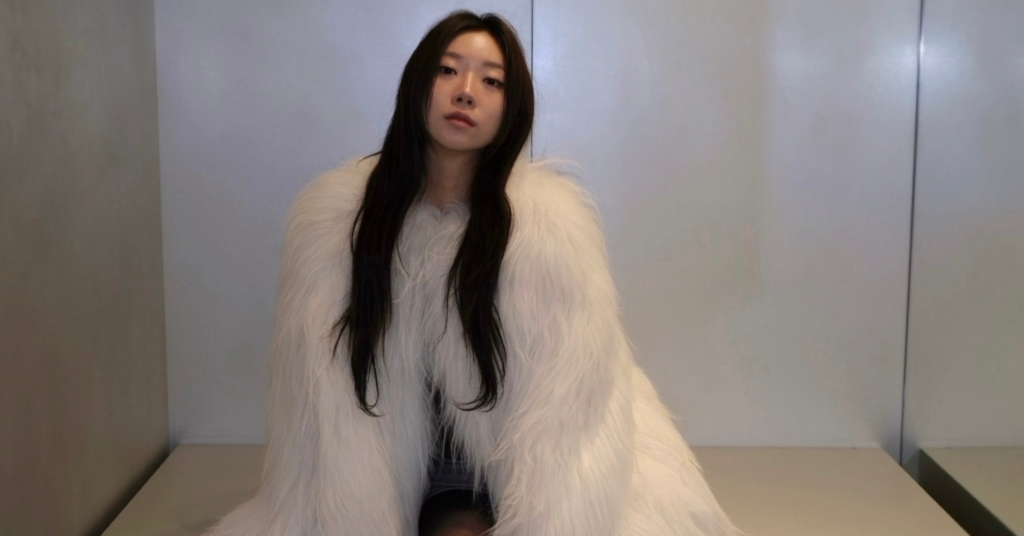 Sujin Gwon posing in a sitting position, wearing a white fur jacket.