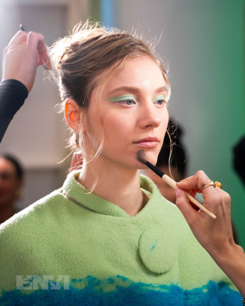 A person with long blonde hair pulled back by someone's hand has makeup applied by a brush to their chin. They wear a light green wool coat with teal whisps below the shoulder and a light green-turquoise eyeshadow.