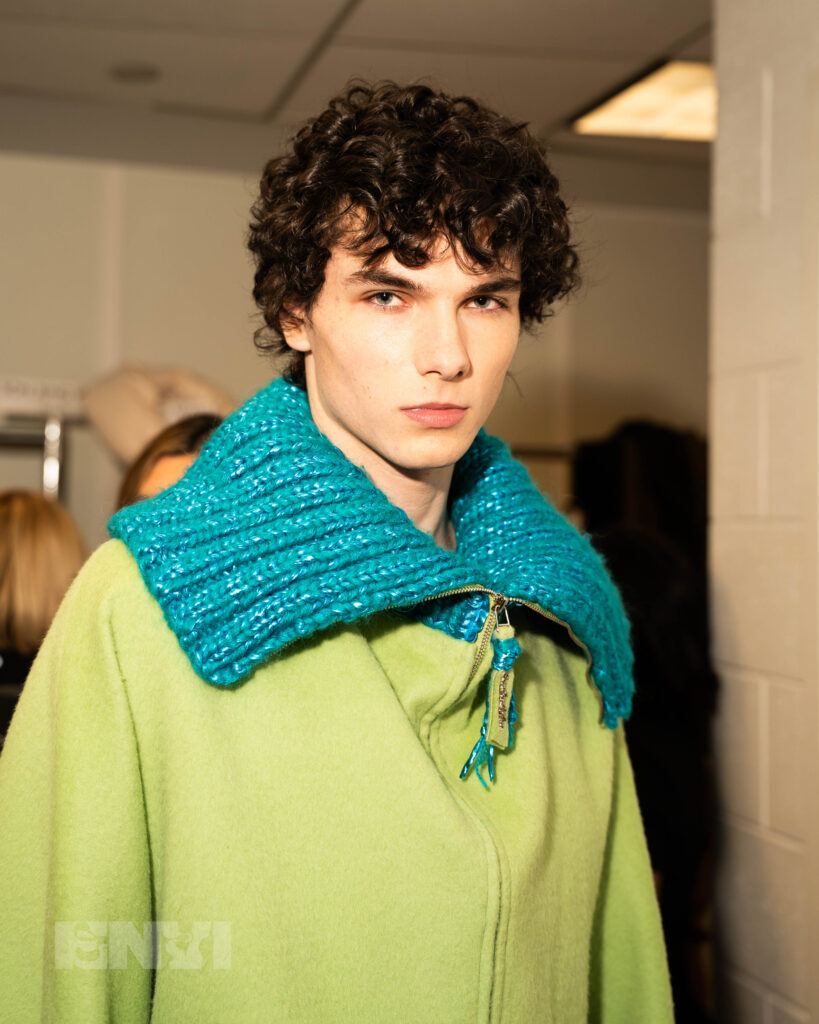 A male model with curly brown hair wears a bright green wool zip-up with teal knitting on the collar and inside.