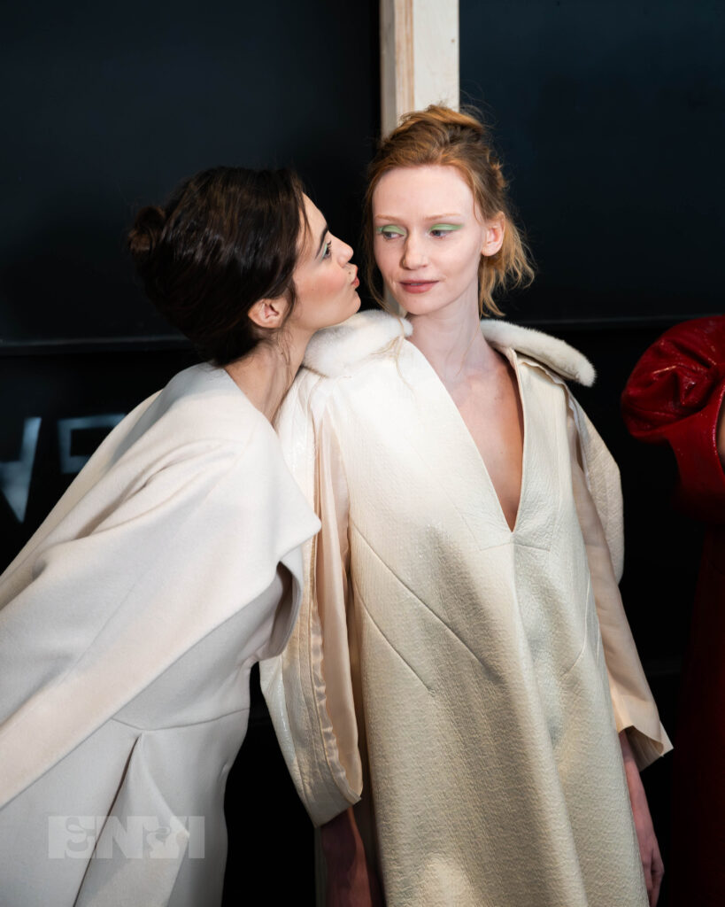 Two models in long white coats pose for a photo. One puckers her lips towards the other that looks at her from the side. They both wear bright green eyeshadow.