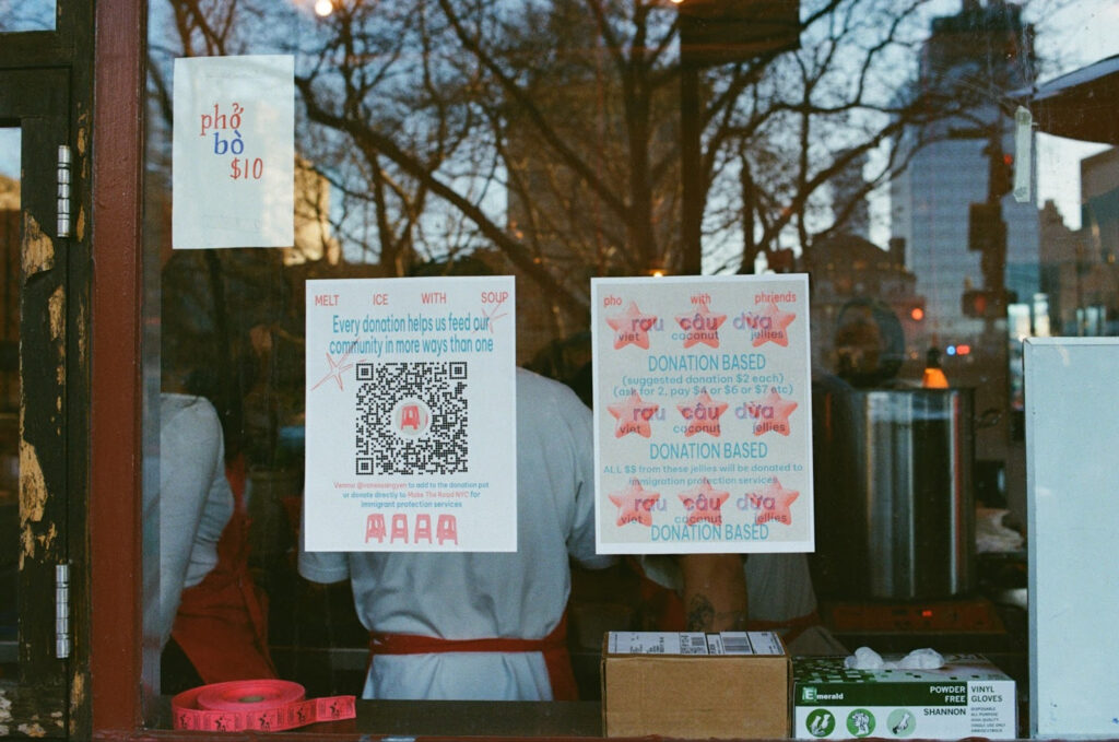 The window to the Phở With Friends popup event at the Time Again Bar, which shows three leaflets pasted to the glass. One says "pho bo $10", one says "Every donation helps us feed our community in more ways than one" and displays a donation QR code, and another says "rou cau dua - viet coconut jellies - donation based (suggested donations $2 each) (ask for 2, pay $4 or $6 or $7 etc)" repeated three times in a row.