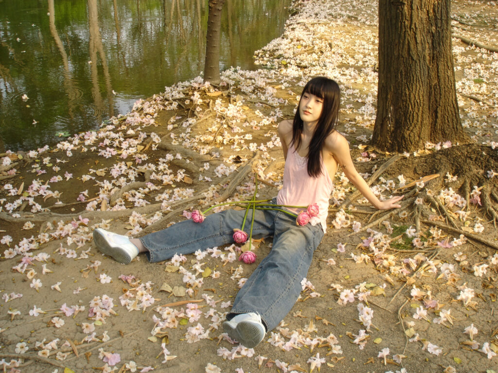 pami sitting under a tree next to a river, the ground around her littered with small pink flowers.
