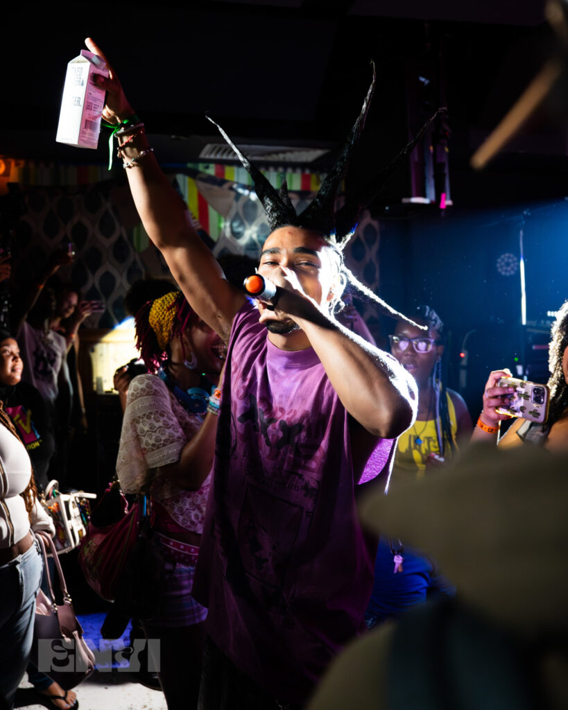 Danny Singh joins the crowd, and sings amongst fans at Baby's All Right. Wearing a purple tank top and spikes in his hair, his left hand hold the mic while his right is lifted into the air holding a box carton drink. 