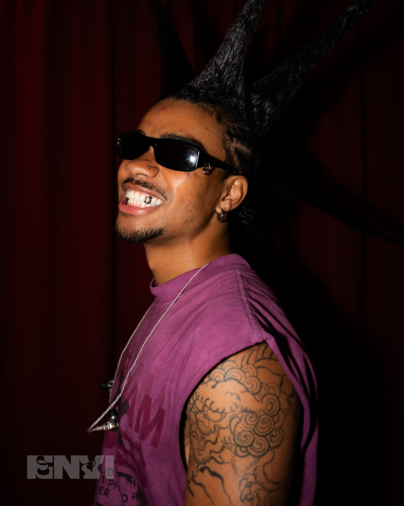 RUOK frontman Danny Singh smiles at the camera with a singular grill on his tooth. Donning spikes in his hair with cornrows he pairs the look with bold sunglasses