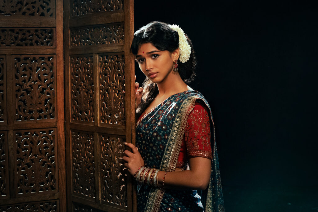"Woman in traditional Indian attire with flowers in her hair stands beside a wooden carved screen in a dimly lit room, looking thoughtfully at the camera."