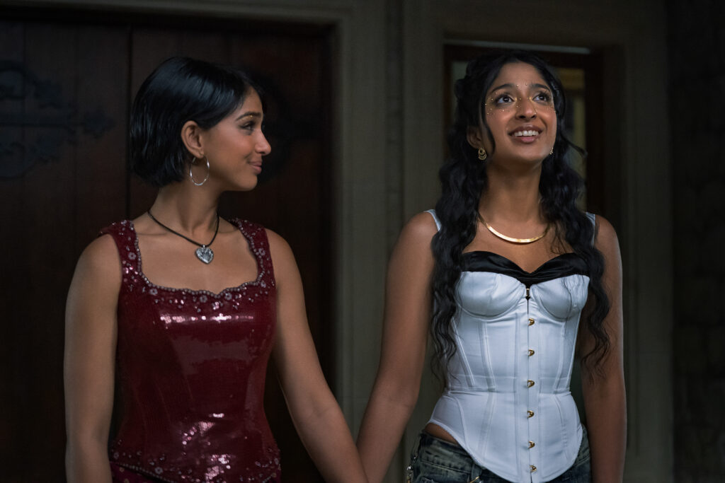 "Two young women dressed up for an event stand indoors, holding hands. One wears a red sequined dress, the other a white corset top. They appear happy and are looking at each other, suggesting friendship or excitement."