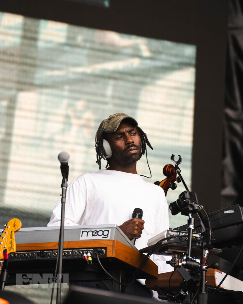 Blood Orange stares out into the distance holding a microphone behind three keyboards.