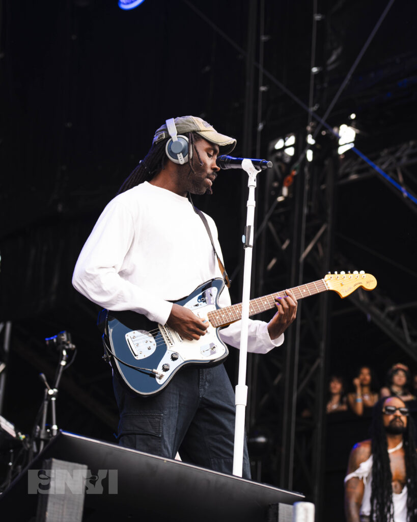 Blood Orange plays a blue and white electric guitar in front of a microphone stand.